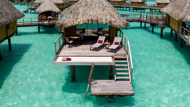 An overwater bungalows with thatched roofs, wooden deck, and lounge chairs.