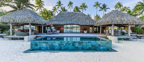 A beachfront resort with thatched-roof cabanas, a swimming pool, and palm trees.