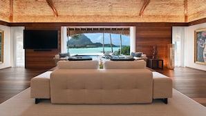 A modern living room with a large sofa, a flat-screen TV, and a view of a tropical beach and mountain.