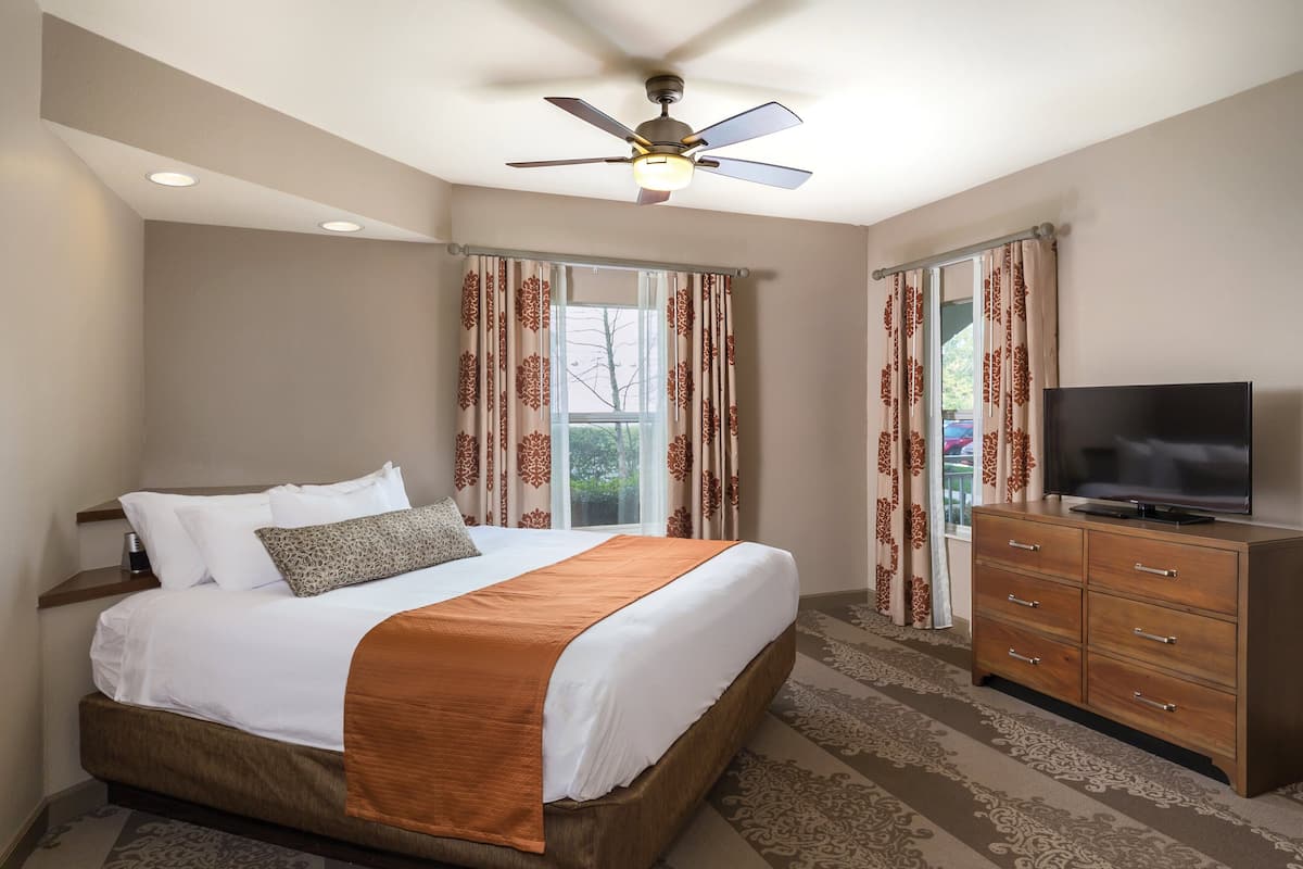 A hotel room with a bed, a TV on a wooden dresser, a ceiling fan, and large windows with curtains.