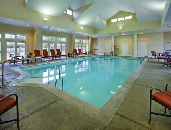 Uma grande piscina interior com várias espreguiçadeiras e mesas, rodeada por amplas janelas e um alto teto.