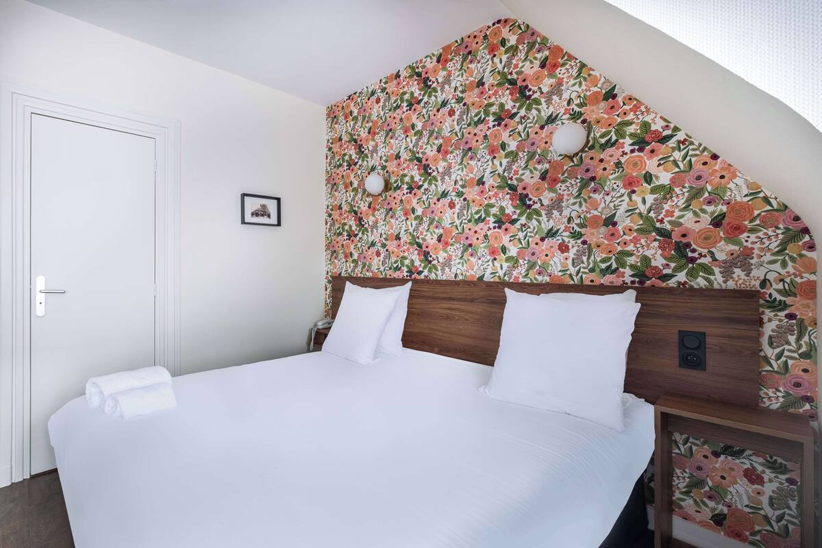 Ein ordentlich bezogenes Bett mit weißen Leinen, ein hölzernes Kopfteil mit floraler Tapete, eine weiße Tür und ein kleines gerahmtes Bild an der Wand.