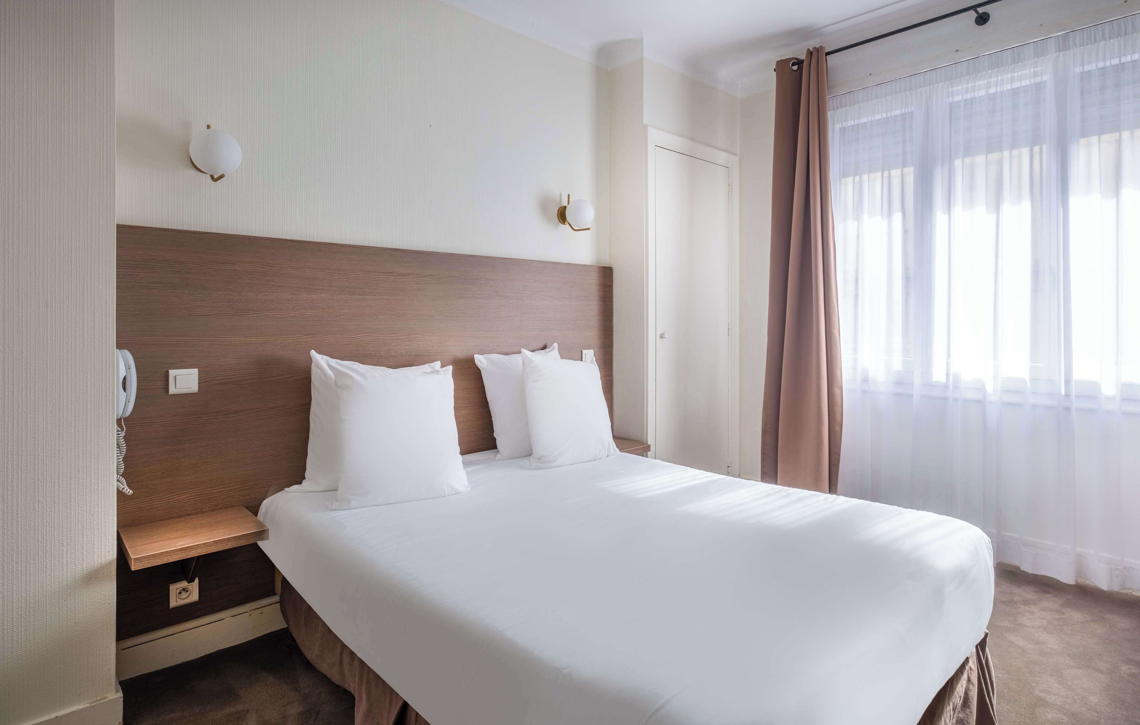A hotel room with a large bed, a wooden headboard, a bedside table with a lamp, and a window with sheer curtains.