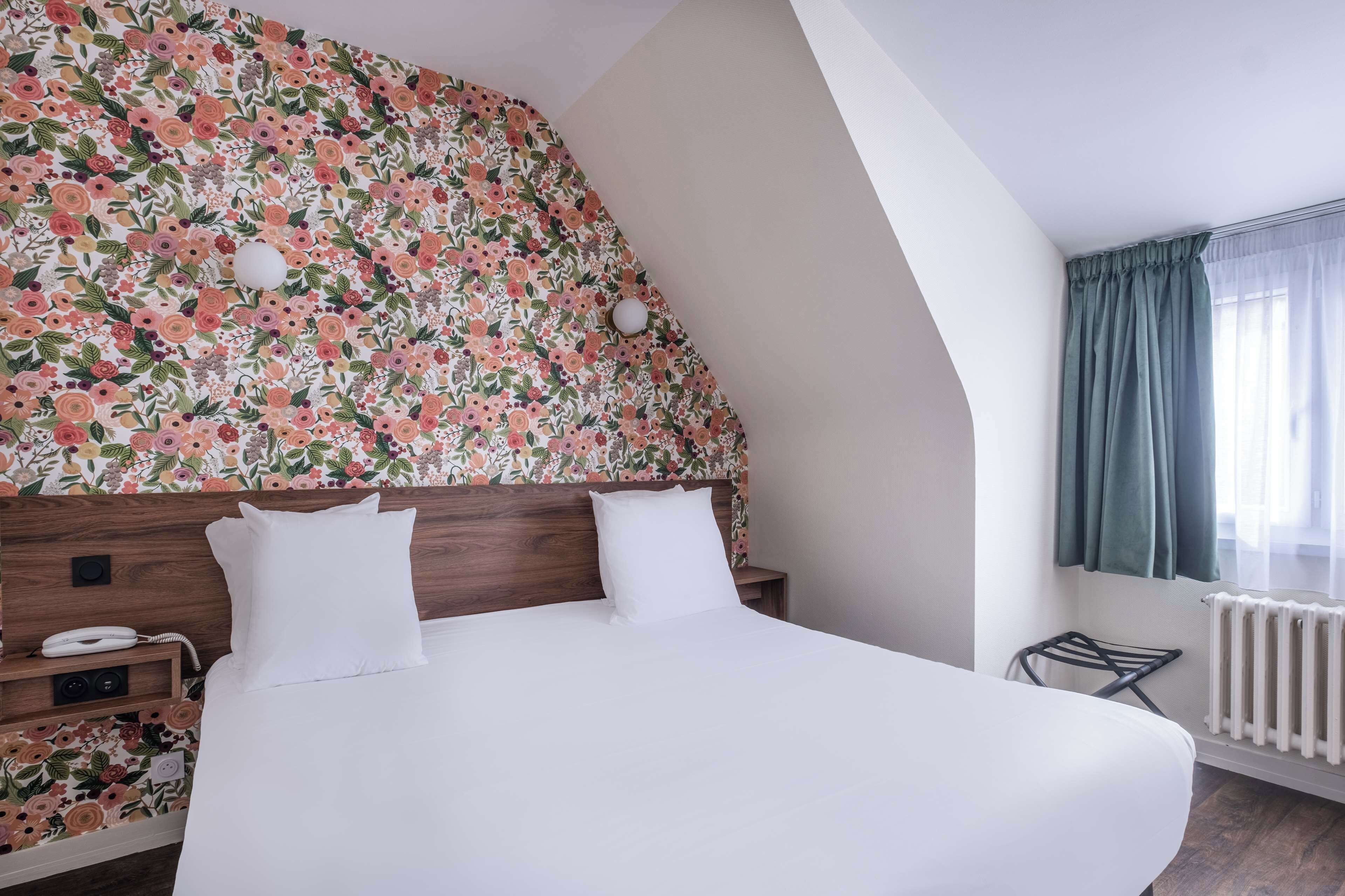 Habitación de hotel con papel tapiz floral, cabecera de madera, cama blanca con dos almohadas, teléfono en la mesita de noche y radiador.
