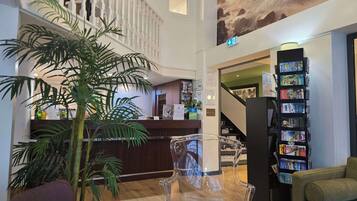 A hotel lobby with a reception desk, a staircase, a bookshelf with magazines, and a large plant.