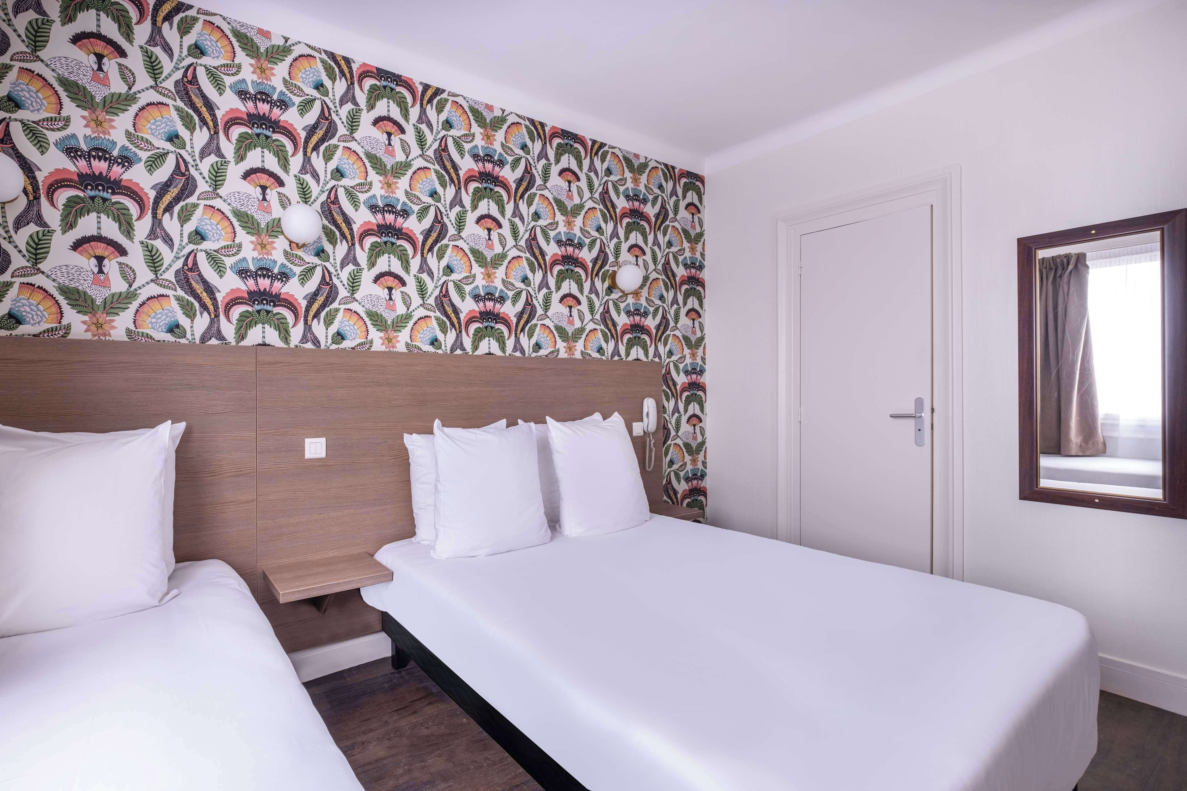 A hotel room with two beds, a patterned wallpaper, a wooden headboard, a bedside table, and a window with curtains.