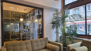 Un interior moderno con un sofá modular, una planta grande en maceta y vistas a un restaurante a través de un separador de vidrio.