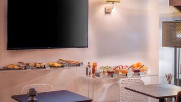 Una habitación de hotel con televisión de pantalla plana, un bufé de desayuno con diversos pasteles, y una zona de estar con mesa y sillas.