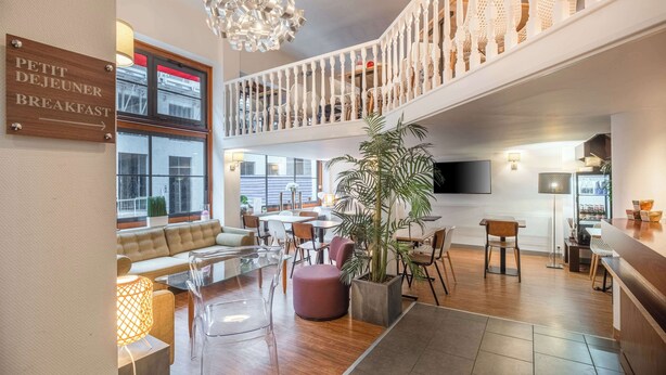 Un moderno vestíbulo de hotel con un cartel que dice 'Petit Dejeuner Breakfast', un área de descanso con sofás y mesas, una escalera a un nivel superior y una planta grande en maceta.