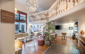 Un moderno vestíbulo de hotel con un cartel que dice 'Petit Dejeuner Breakfast', un área de descanso con sofás y mesas, una escalera a un nivel superior y una planta grande en maceta.