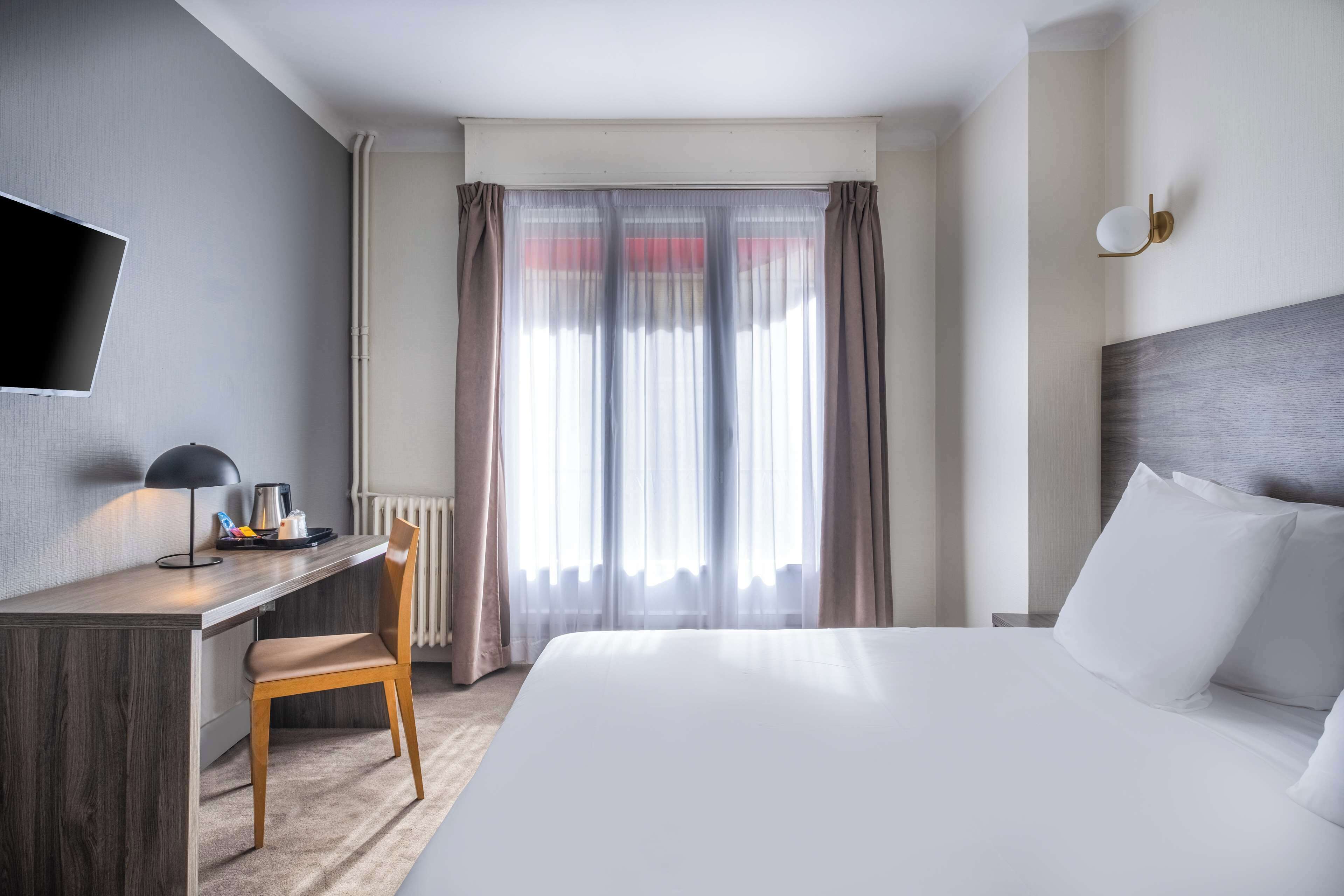 Habitación de hotel con cama, escritorio, silla, televisor y ventana con cortinas.