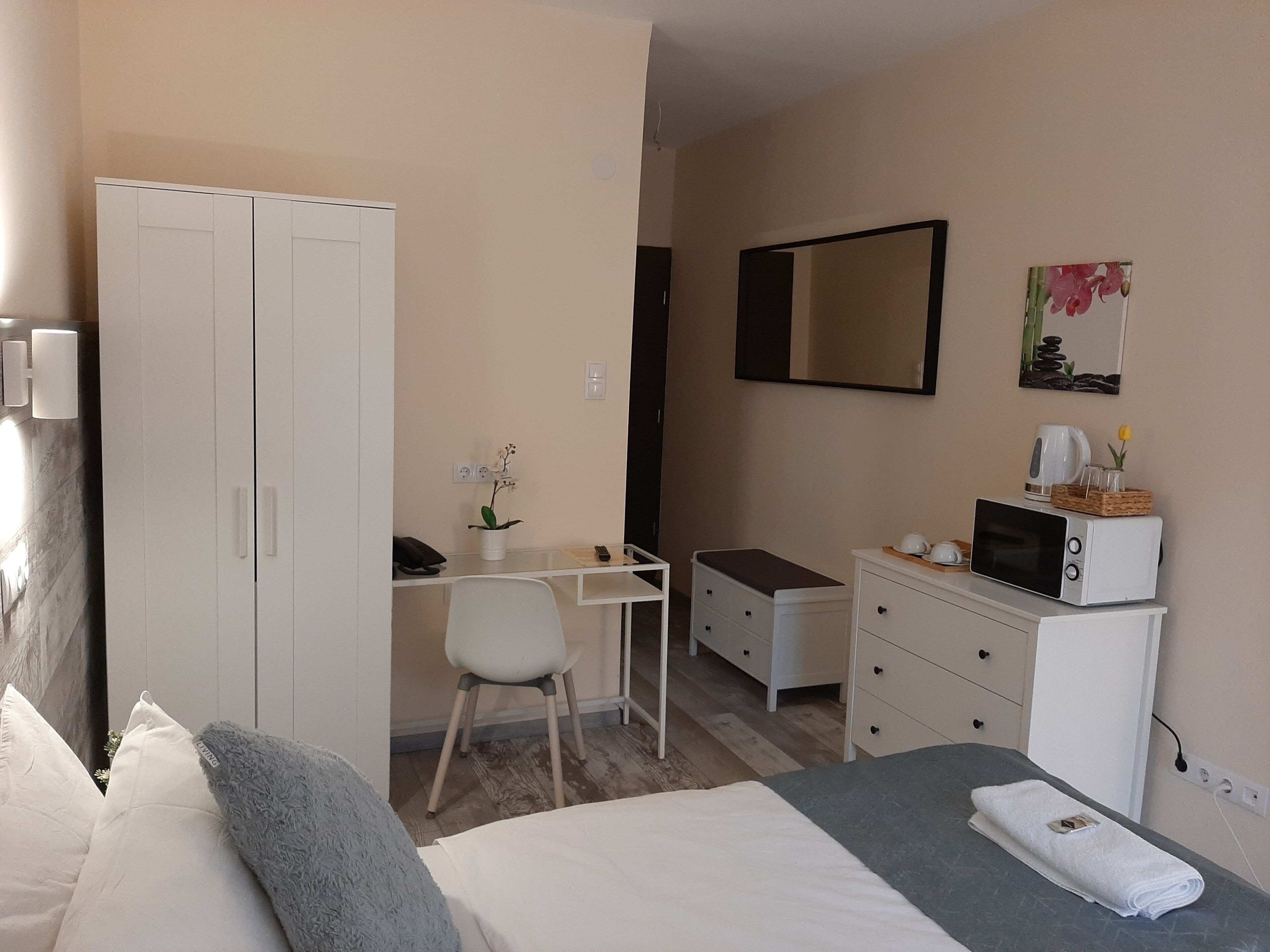A hotel room with a bed, a desk, a chair, a wardrobe, a dresser, a microwave, and a small table with a plant.