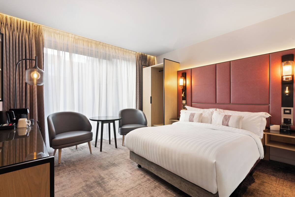 Habitación de hotel con una cama grande, dos sillones, un escritorio con cafetera y vistas al exterior a través de cortinas translúcidas.