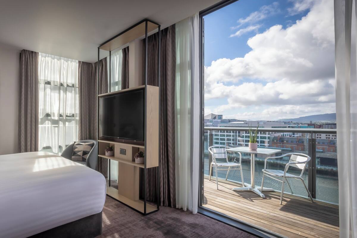 Habitación de hotel con cama, televisión sobre un soporte de madera, balcón con sillas y vista a la ciudad.