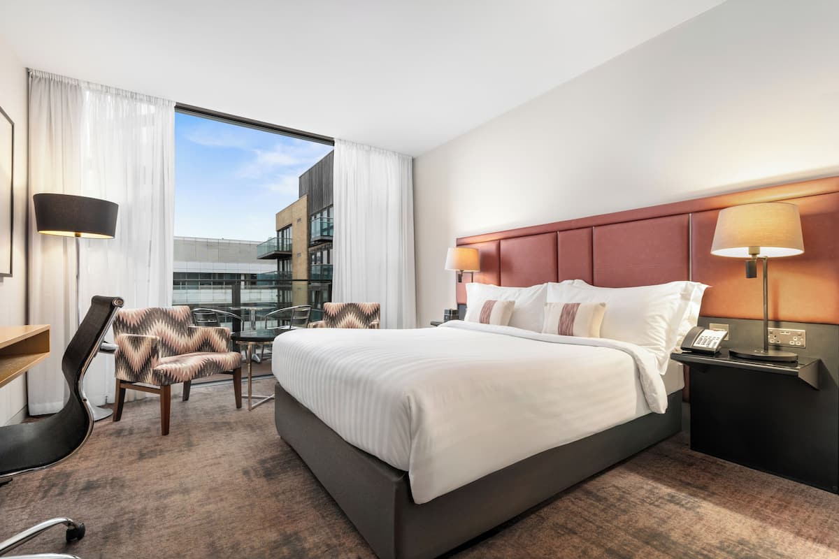 A hotel room with a large bed, a desk, a chair, a lamp, and a view of a building.