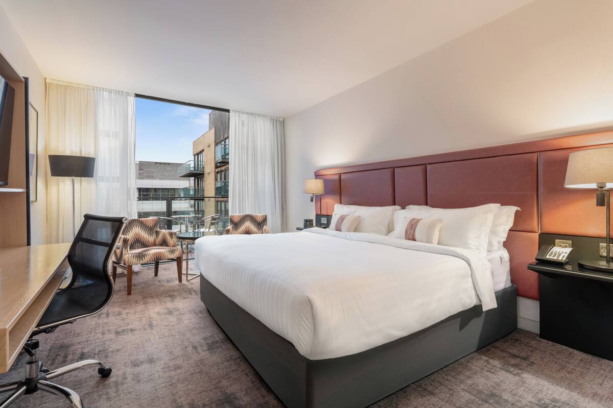 Habitación de hotel con una cama grande, un escritorio, una silla, una lámpara y vistas a un edificio.