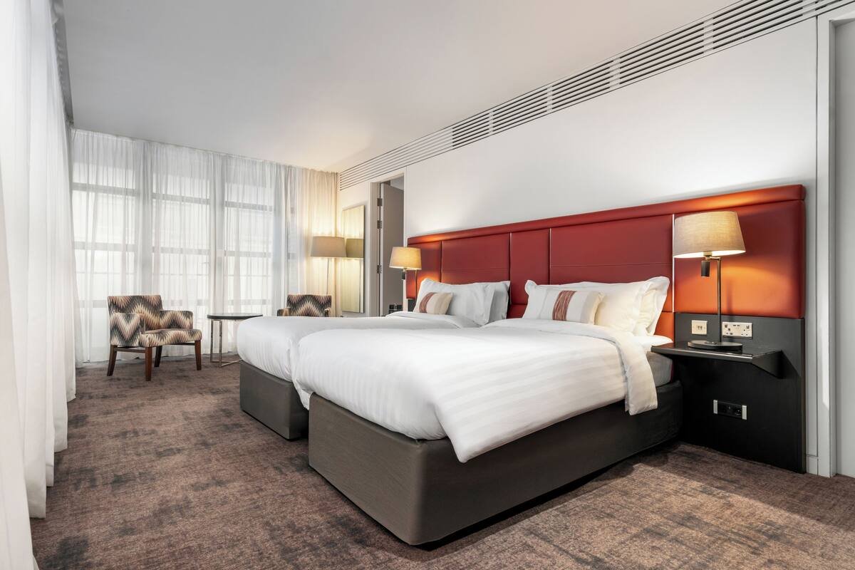Una habitación de hotel con una cama grande, dos mesitas de noche, una silla y un ventanal con cortinas.