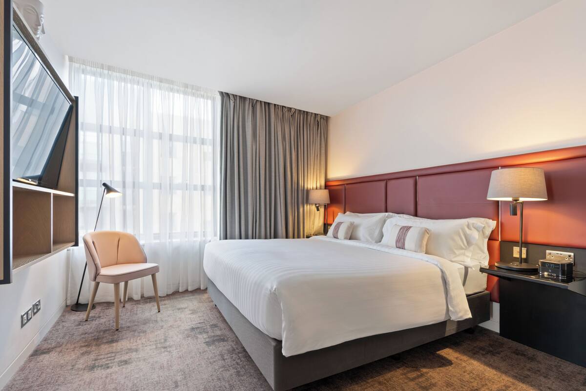 Una habitación de hotel con una cama grande, una silla, un escritorio con lámpara y un televisor montado en la pared.