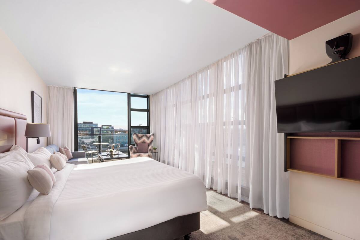 Habitación de hotel moderna con una cama grande, un televisor, un balcón con vista a la ciudad y un ventanal con cortinas traslúcidas.
