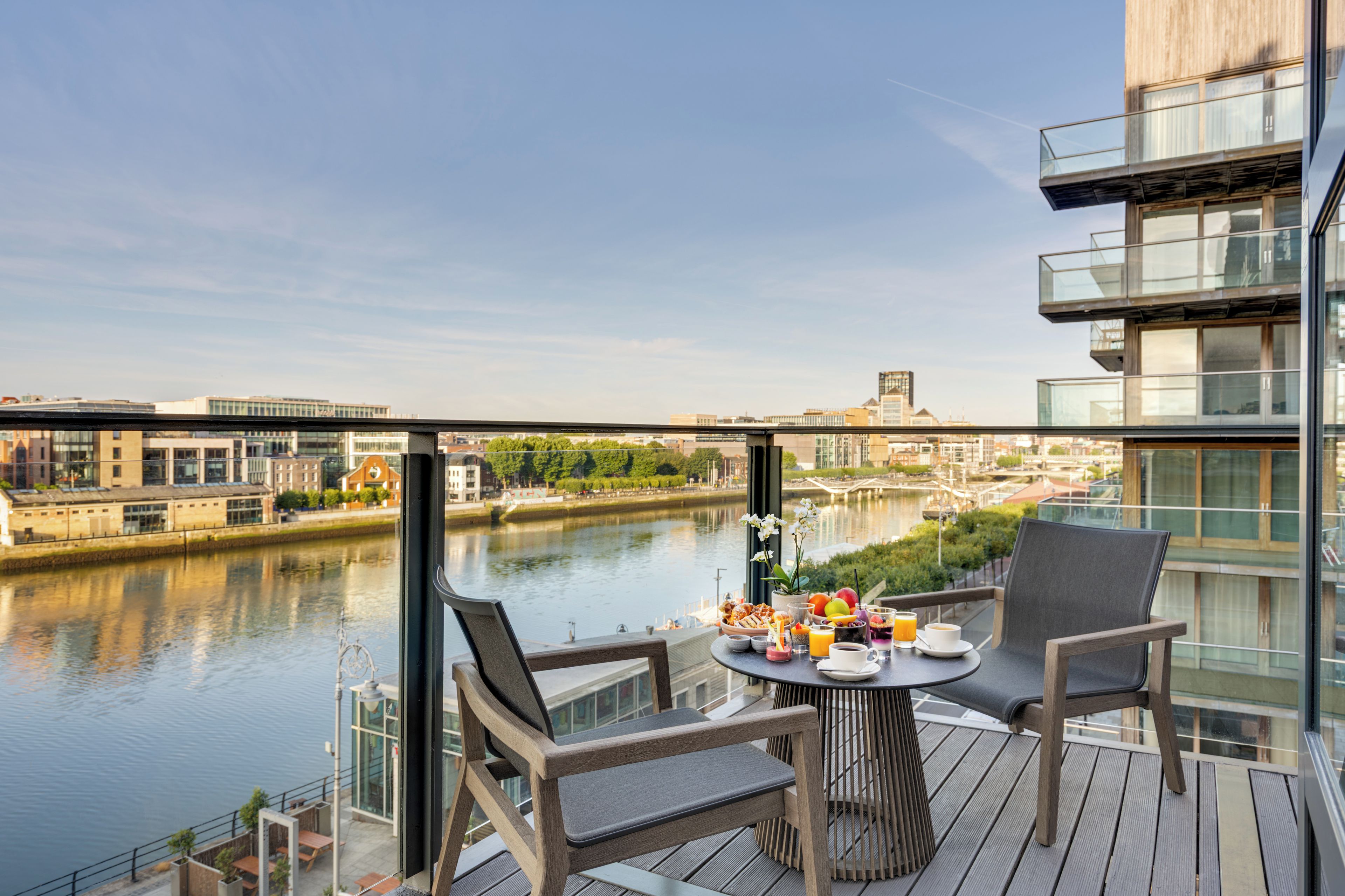 A balcony with a table set for breakfast overlooking a river, with modern buildings in the background.