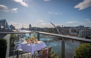 Una mesa con mantel blanco, un vaso de jugo de naranja y un vaso de agua en un balcón con vista a un paisaje urbano con edificios modernos y un puente.