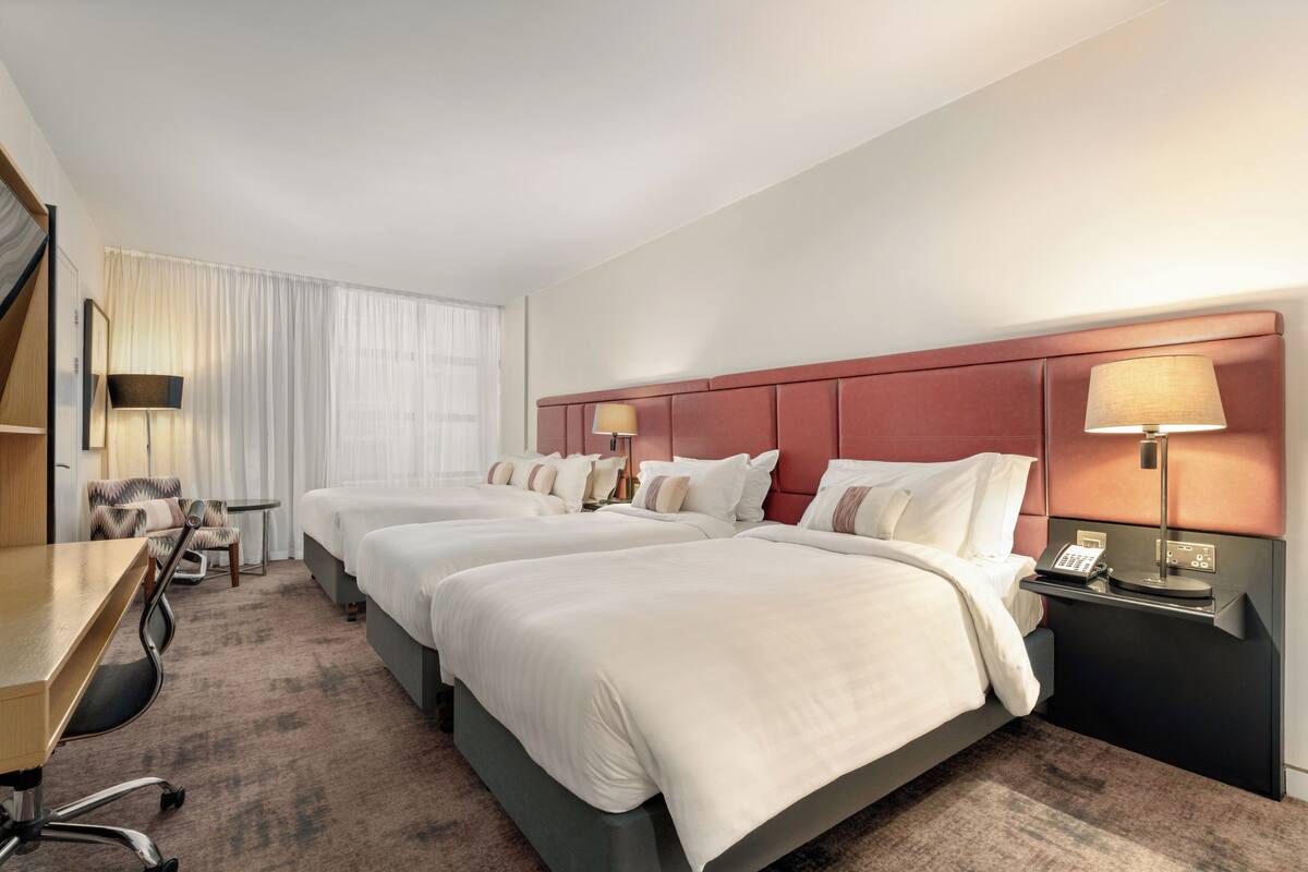Habitación de hotel con tres camas, un escritorio, una silla, una lámpara y un teléfono en la mesita de noche.