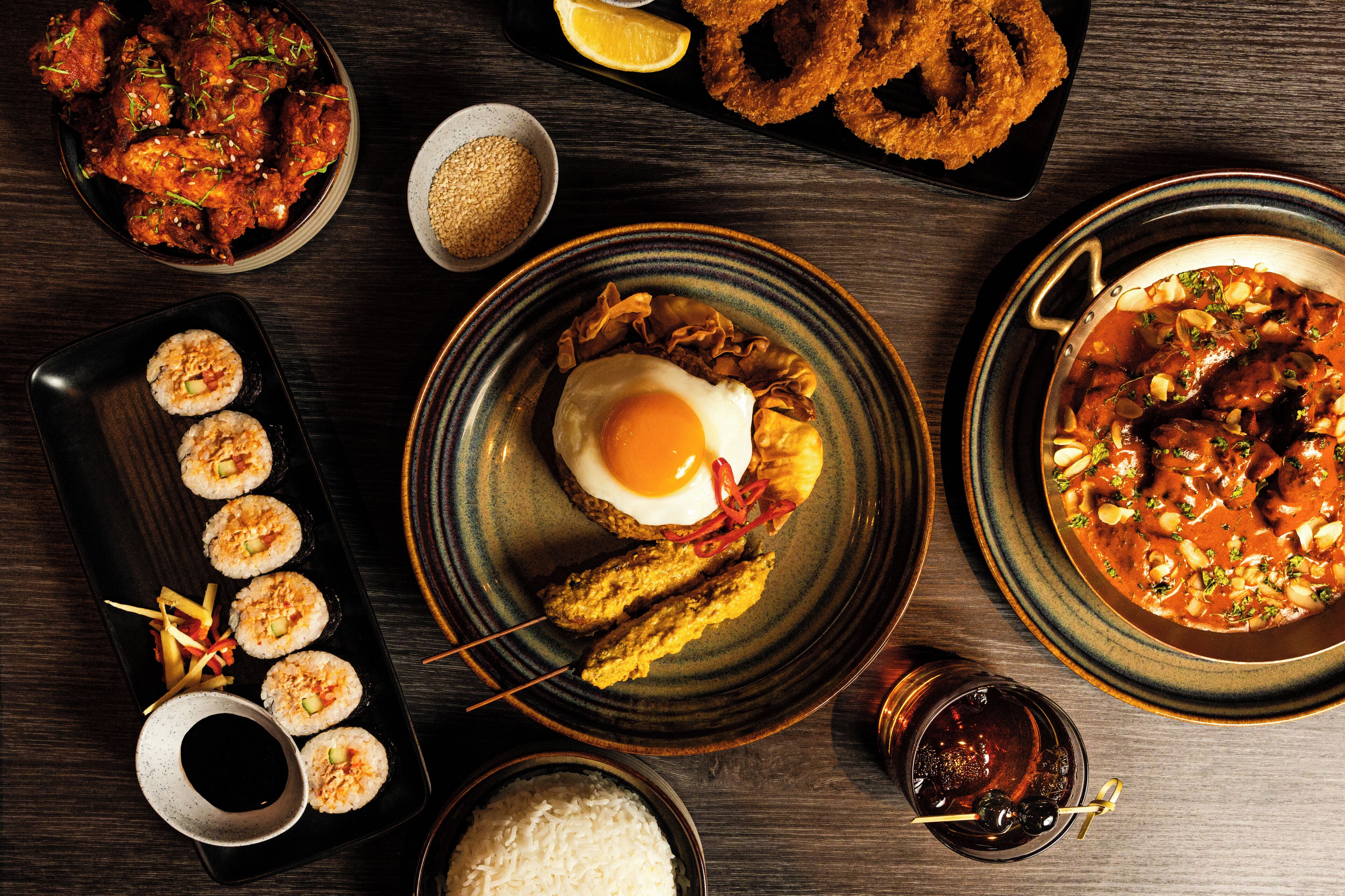 A variety of dishes including fried chicken, sushi, and a bowl of soup, arranged on a wooden table.