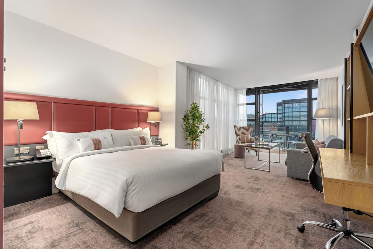 Habitación de hotel con una cama grande, un escritorio con silla, una mesita, una planta y vistas a un edificio a través de la ventana.