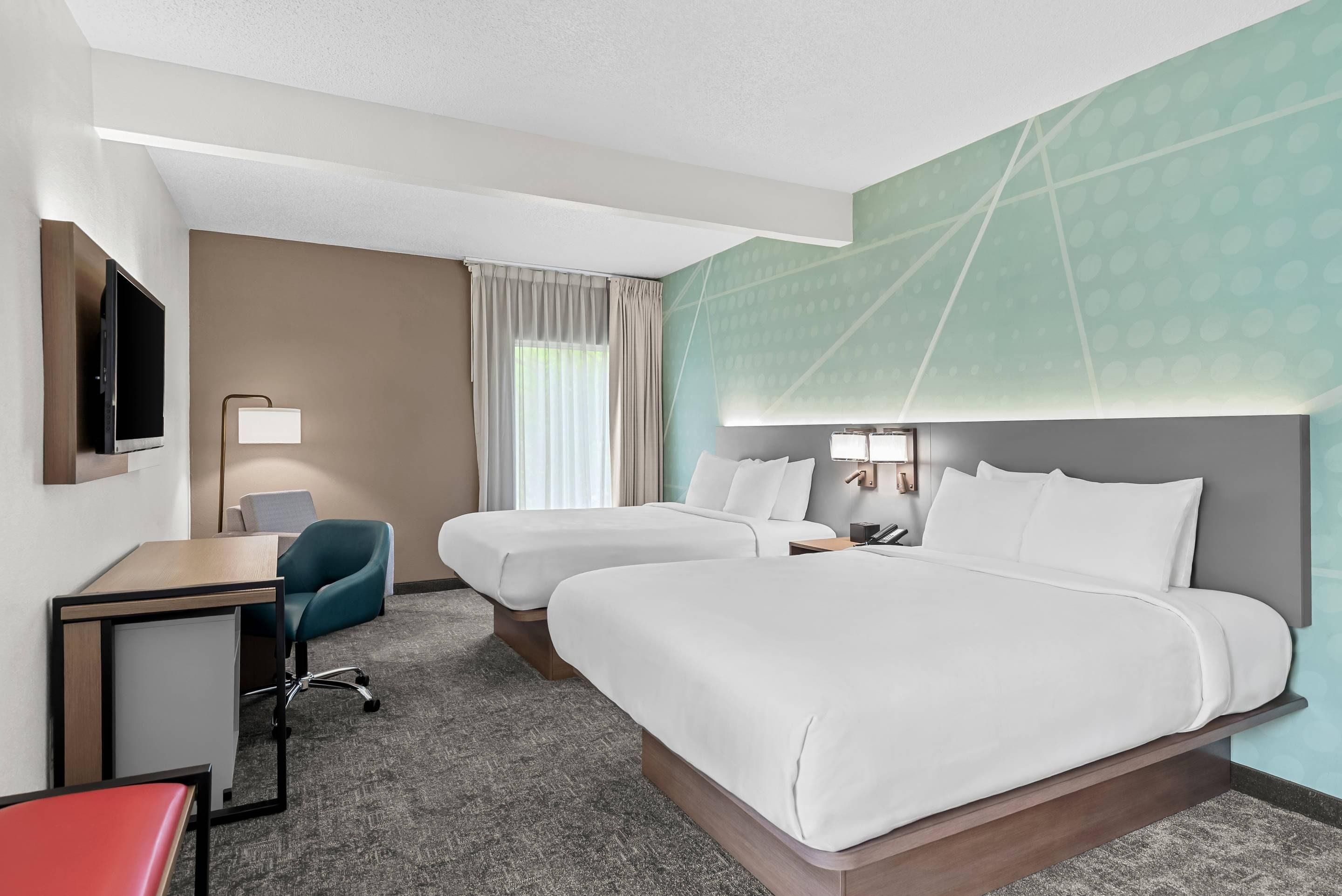 A hotel room with two beds, a desk, a chair, a TV, and a window with curtains.