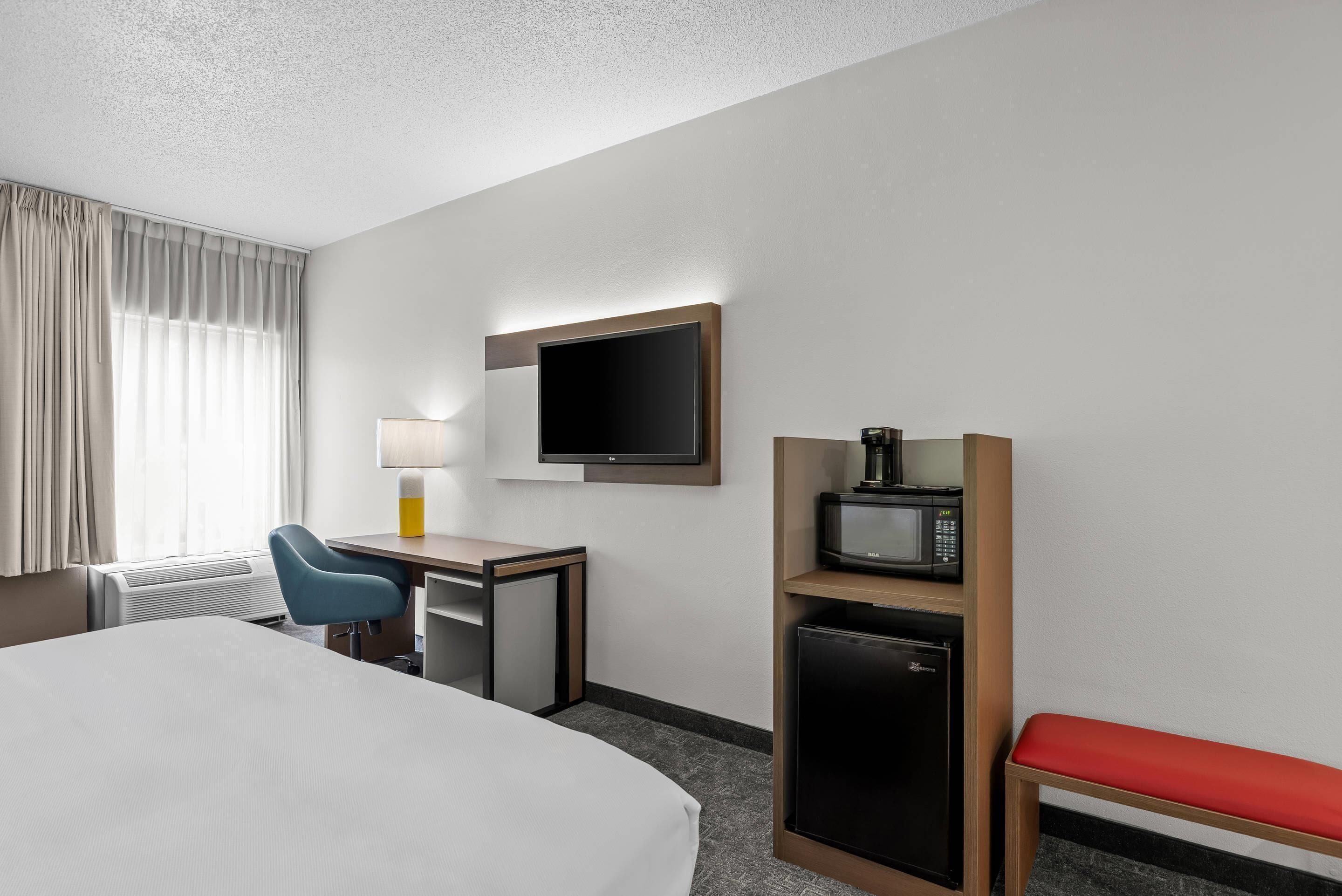 Ett hotellrum med en säng, ett skrivbord, en stol, en TV, en mikrovågsugn, ett litet kylskåp och en röd bänk.