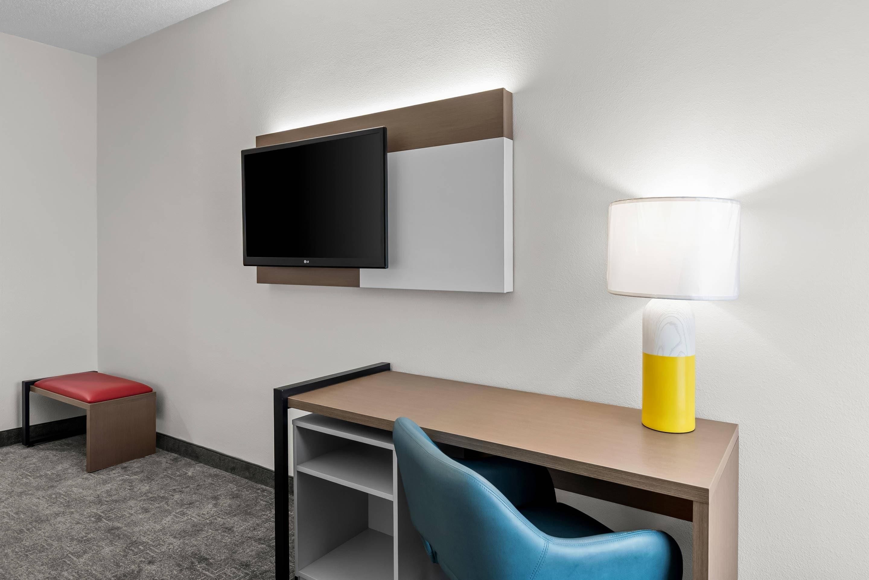 Habitación de hotel con televisión de pantalla plana, escritorio con silla, taburete pequeño y lámpara de noche.