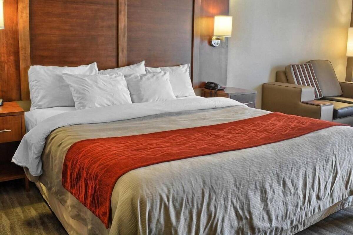 A hotel room with a large bed, a wooden headboard, a nightstand, a desk, a chair, and a wall-mounted lamp.
