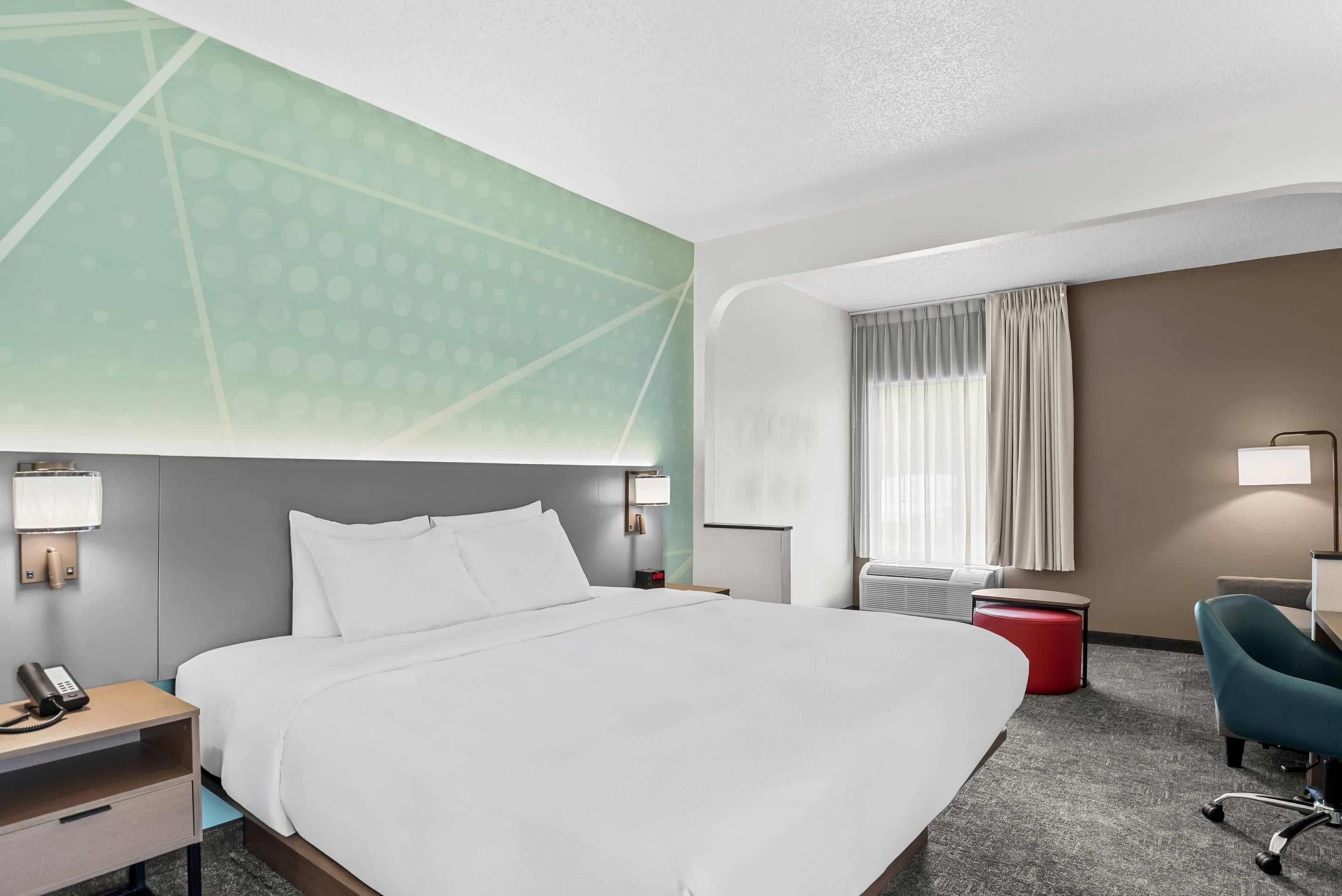 Habitación de hotel con una cama grande, un escritorio con teléfono, un reposapiés rojo, una silla azul y un aire acondicionado instalado en la pared.
