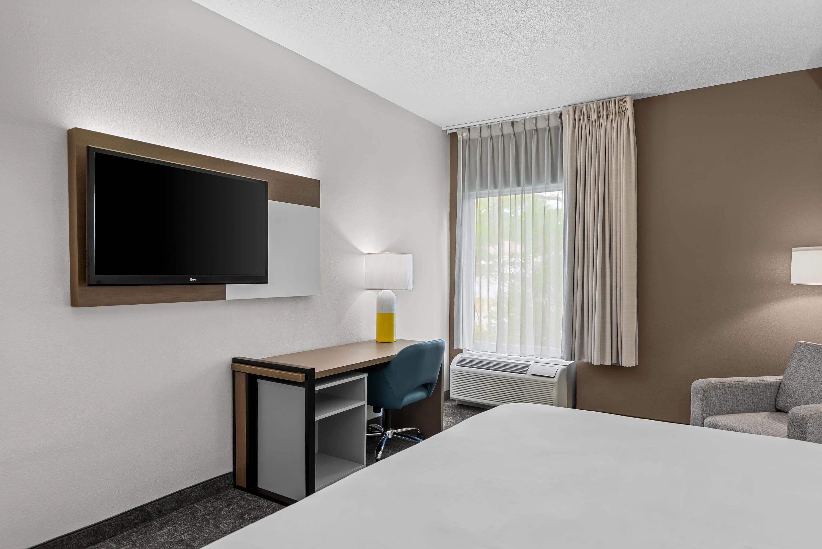 A hotel room with a bed, a desk with a chair, a TV, a lamp, a window with curtains, and an air conditioning unit.
