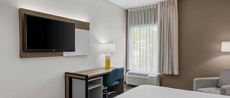 A hotel room with a bed, a desk with a chair, a TV, a lamp, a window with curtains, and an air conditioning unit.
