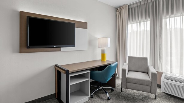 A hotel room with a flat-screen TV mounted on the wall, a desk with a chair, a lamp, and a window with curtains. - Comfort Inn Ruther Glen near Kings Dominion (Ruther Glen)