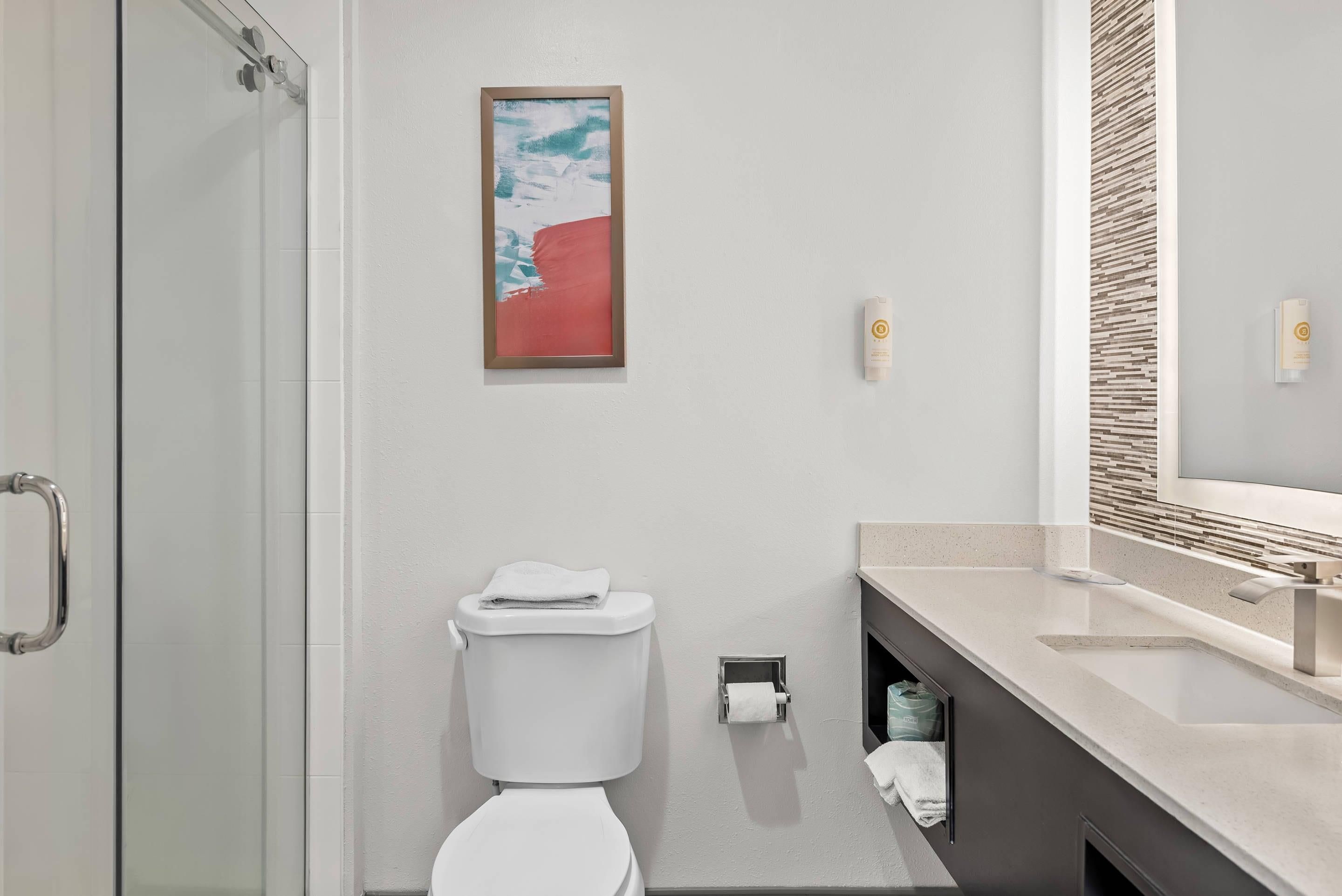 A modern bathroom with a glass shower enclosure, a toilet with a towel on top, a framed artwork on the wall, a sink with a mirror, and a towel dispenser.