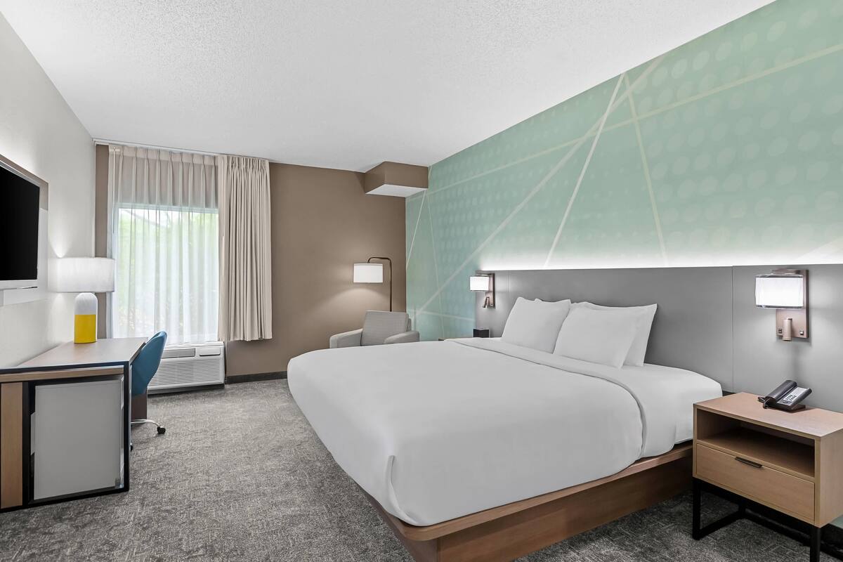 A hotel room with a bed, desk, chair, bedside table, lamp, and a large window with curtains.