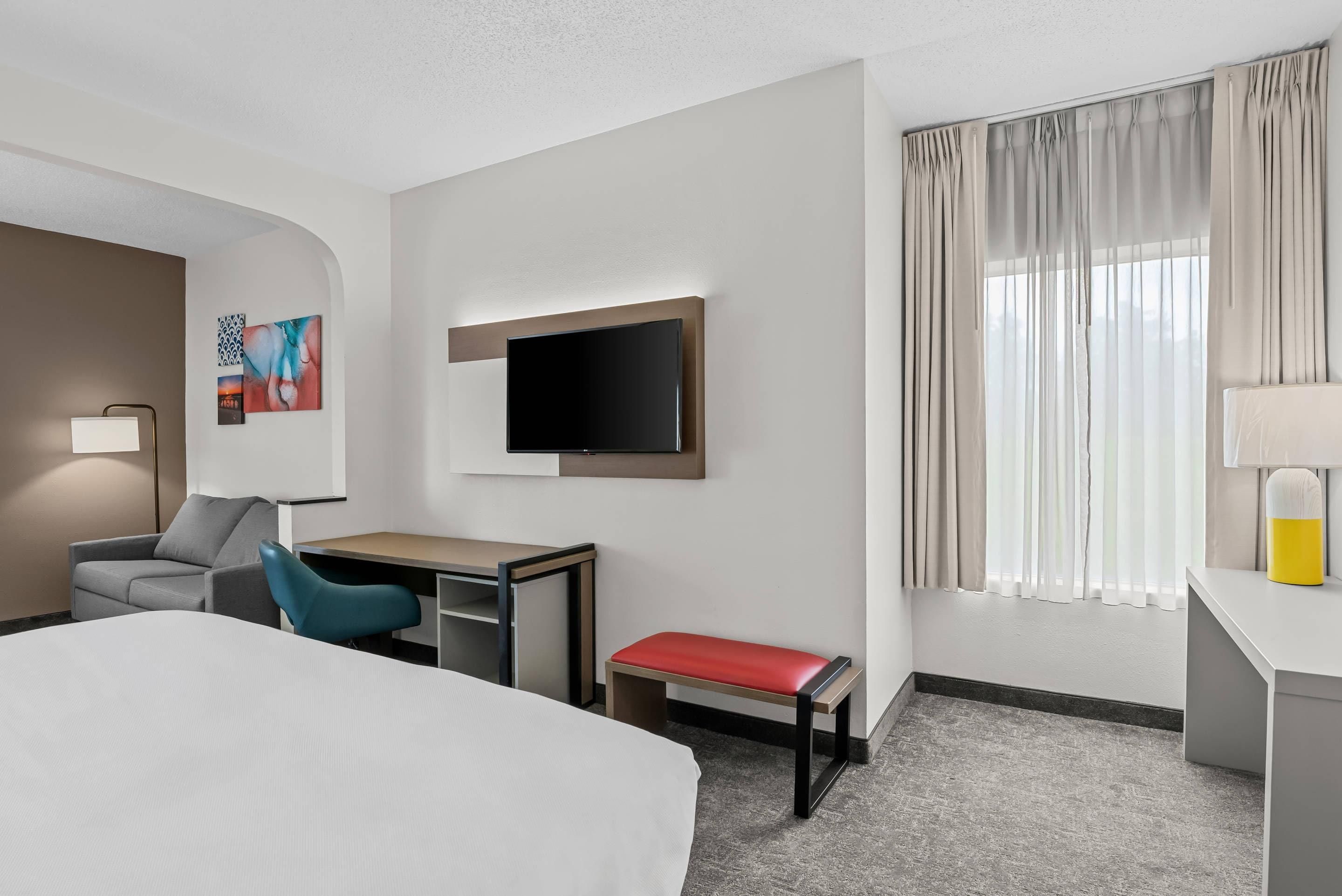 Une chambre d’hôtel avec un lit, un bureau, une chaise, une télévision et une fenêtre avec des rideaux.