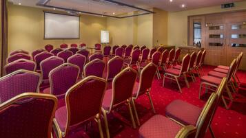 Un salón de conferencias con hileras de sillas rojas, una pantalla de proyector y una pizarra.
