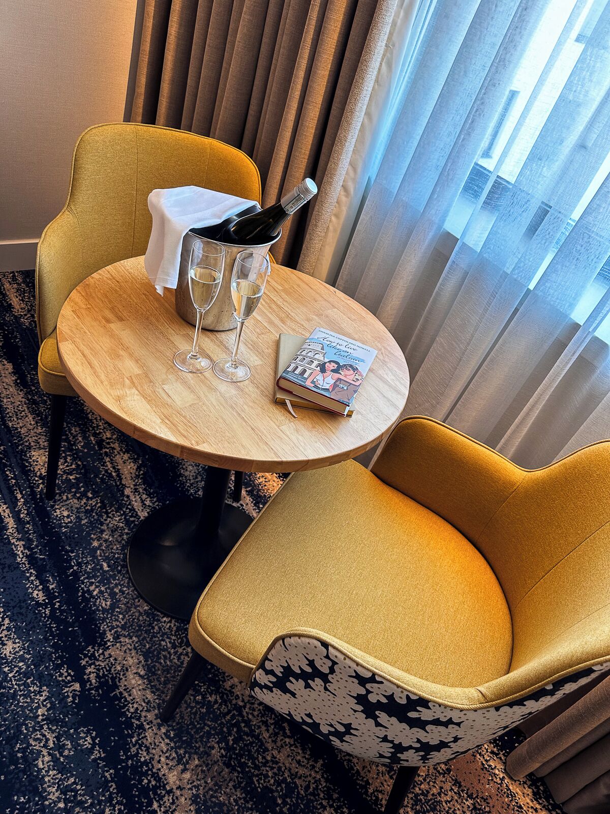 A hotel room with a round wooden table, two yellow armchairs, a bottle of champagne, two glasses, and a book.