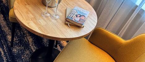 A hotel room with a round wooden table, two yellow armchairs, a bottle of champagne, two glasses, and a book.