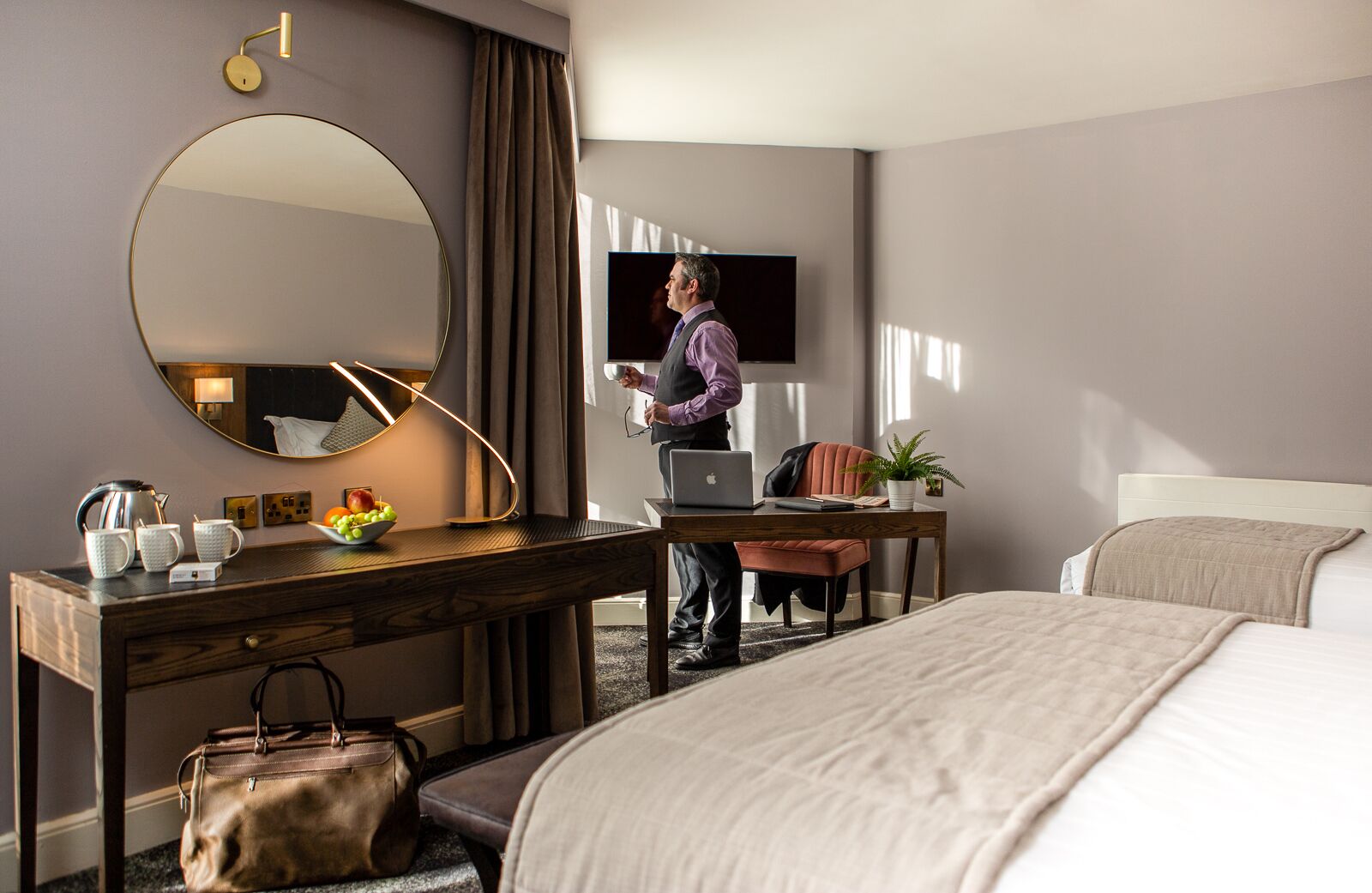 A hotel room with a bed, a desk with a laptop, a mirror, a television, and a potted plant.