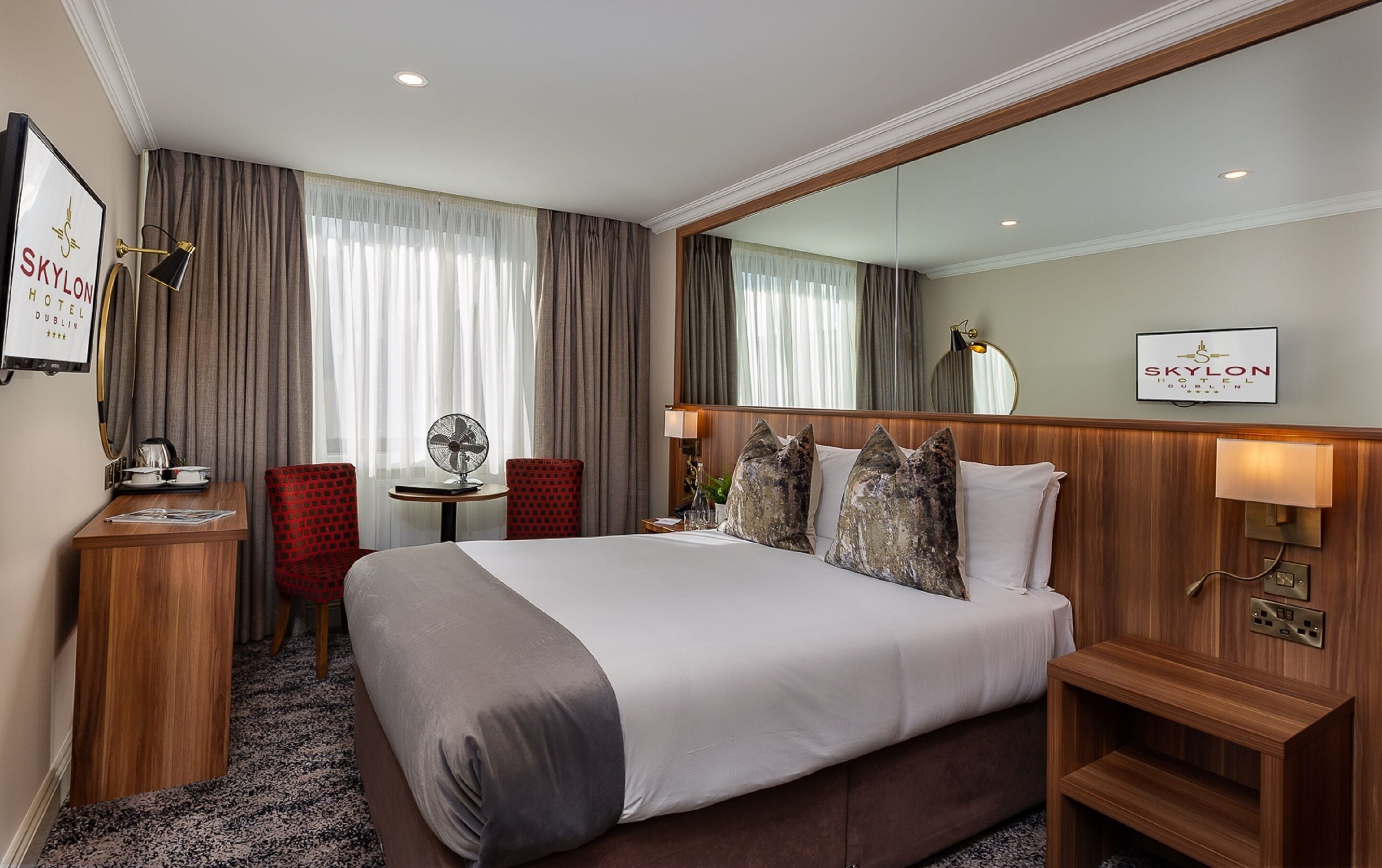 Une chambre d  ’hôtel avec un grand lit, un bureau avec une chaise, une petite table avec un ventilateur et un miroir au mur.