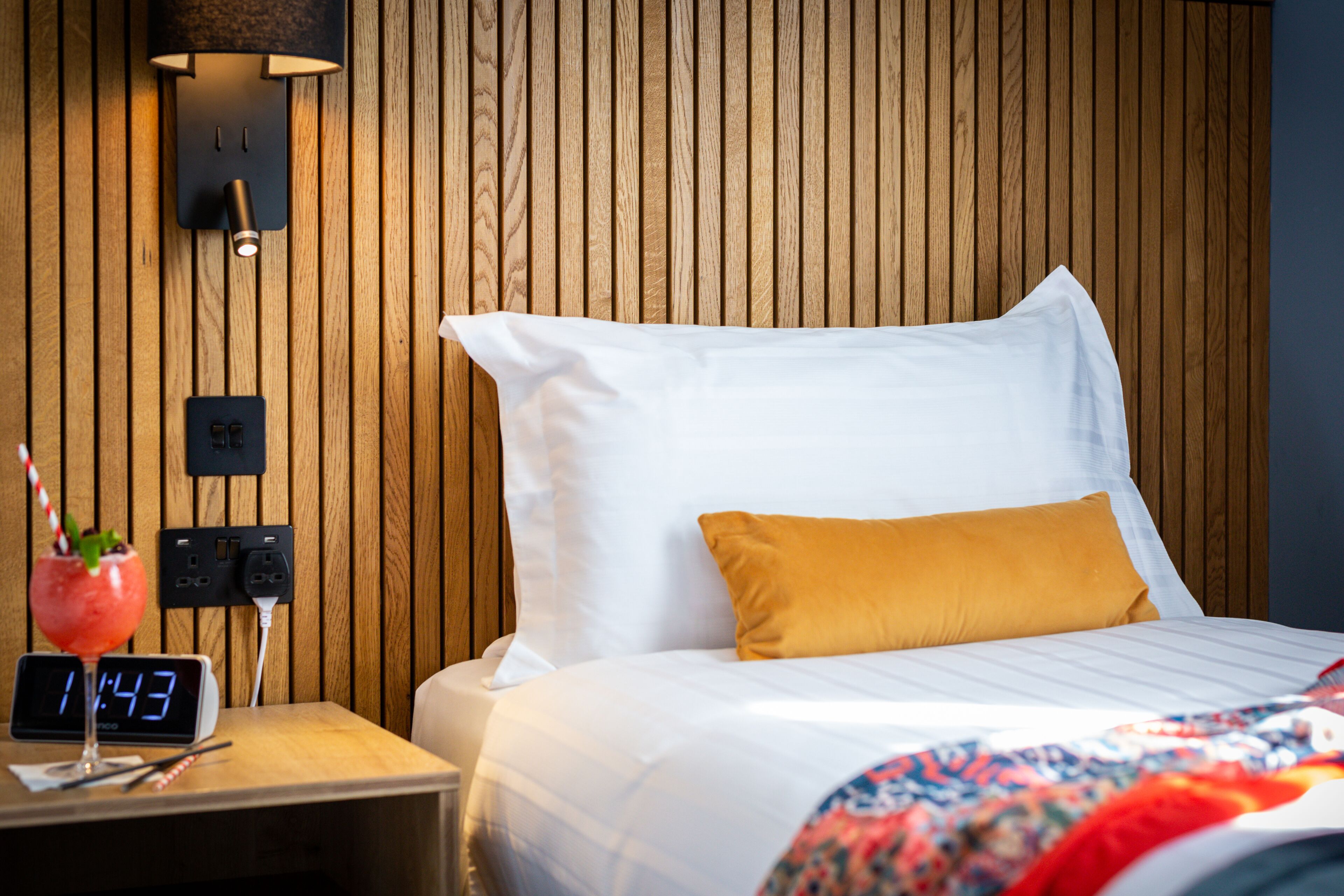 A neatly made bed with white and yellow pillows, a wooden headboard, a bedside table with a clock, and a wall-mounted lamp.
