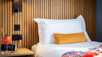 A neatly made bed with white and yellow pillows, a wooden headboard, a bedside table with a clock, and a wall-mounted lamp.