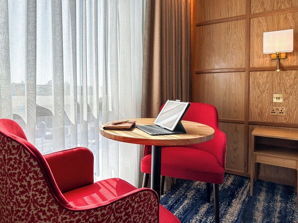Habitación de hotel con una mesa redonda de madera, sillas tapizadas en rojo, una computadora portátil, una libreta y un bolígrafo.