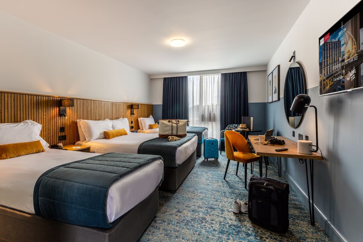 Habitación de hotel con dos camas, un escritorio, una silla, una maleta y un espejo.