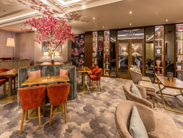 Un moderno vestíbulo de hotel con una zona de estar central que incluye sillones cómodos y una mesa de madera. Un llamativo arreglo floral se exhibe en el centro.