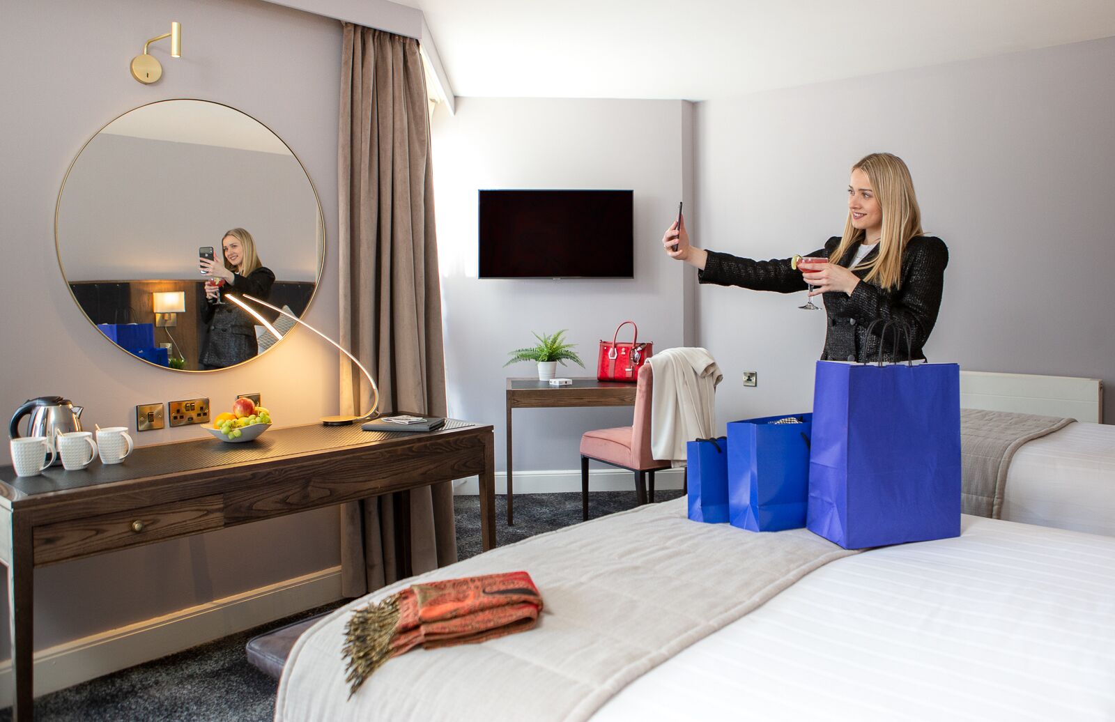 A hotel room with a bed, a desk with a lamp, a mirror, a TV, and a red handbag on a chair.